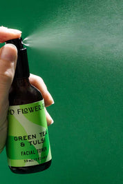 A hand sprays a mist from a brown bottle labeled Good Flower Farm Green Tea & Tulsi Facial Toner against a solid green background.