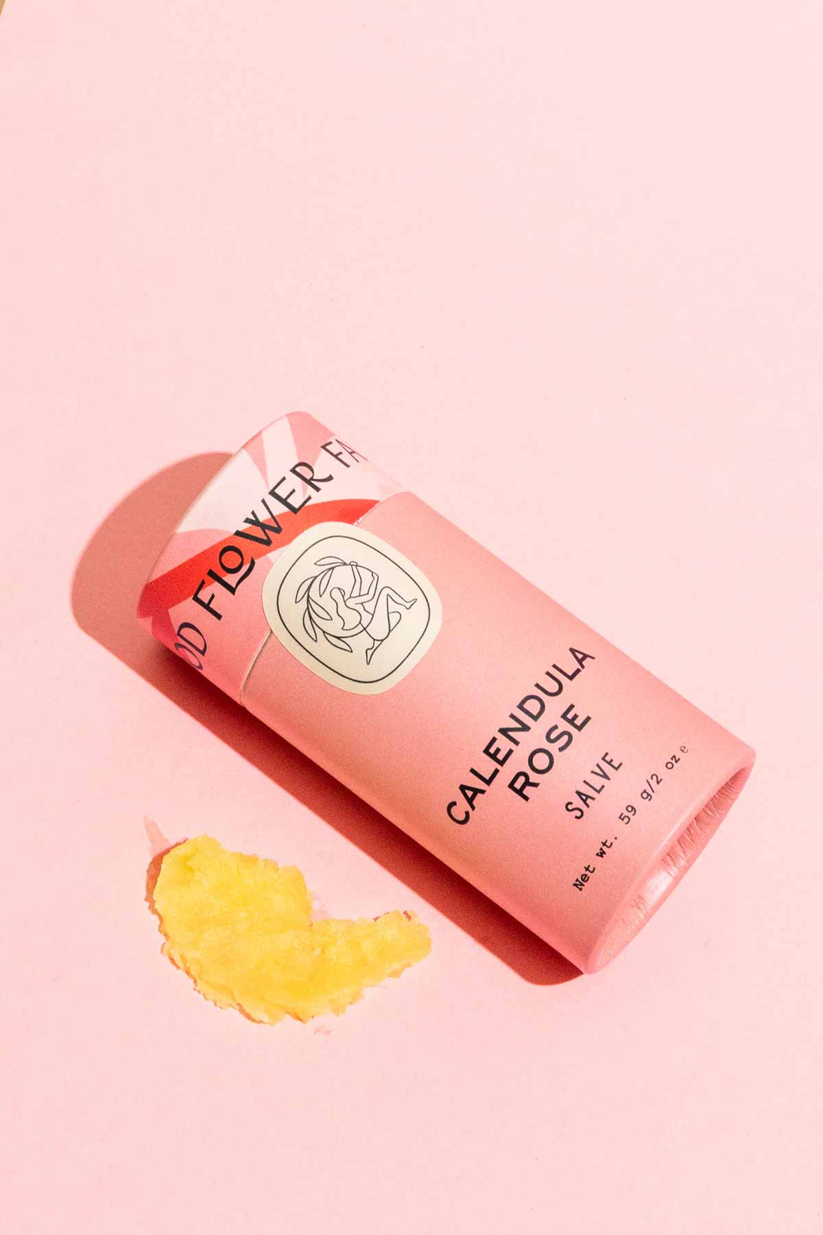 A pink tube of Good Flower Farm Calendula Rose Salve rests on a pink background with a dab of yellow herbal salve nearby. The eco-friendly packaging features a simple floral illustration.