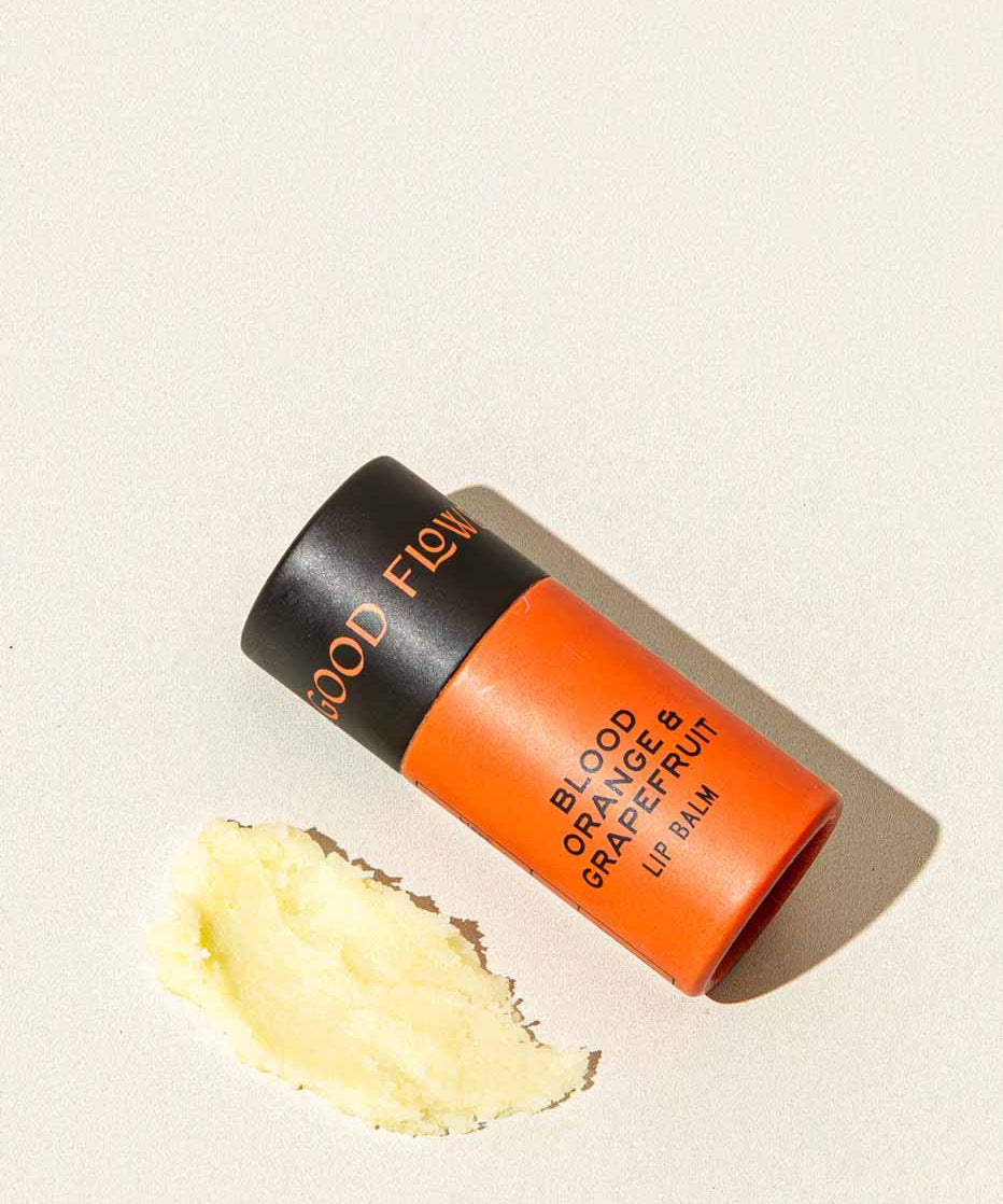 A tube of Good Flower Farm Blood Orange & Grapefruit Organic Lip Balm in orange and black rests on a light surface, beside a small smear of the moisturizing, eco-friendly balm.