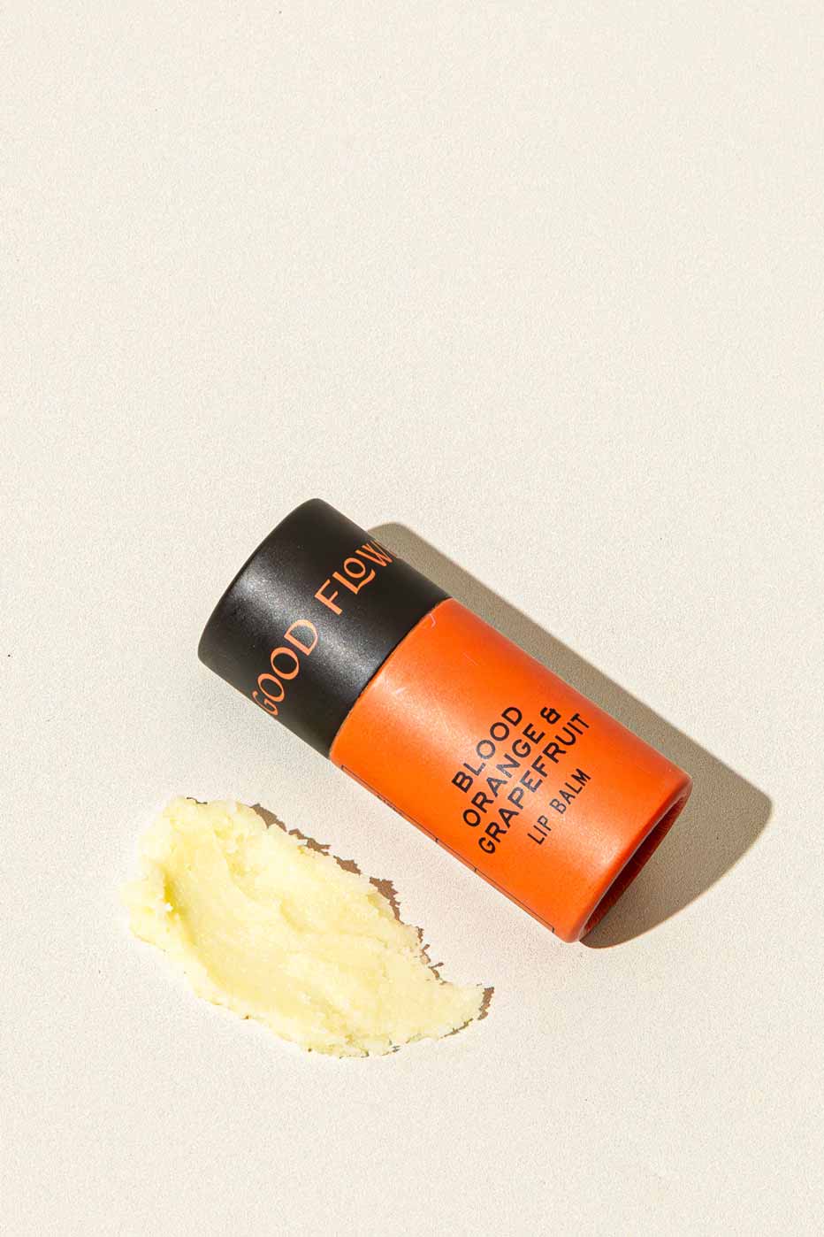 A tube of Good Flower Farm Blood Orange & Grapefruit Organic Lip Balm in orange and black rests on a light surface, beside a small smear of the moisturizing, eco-friendly balm.