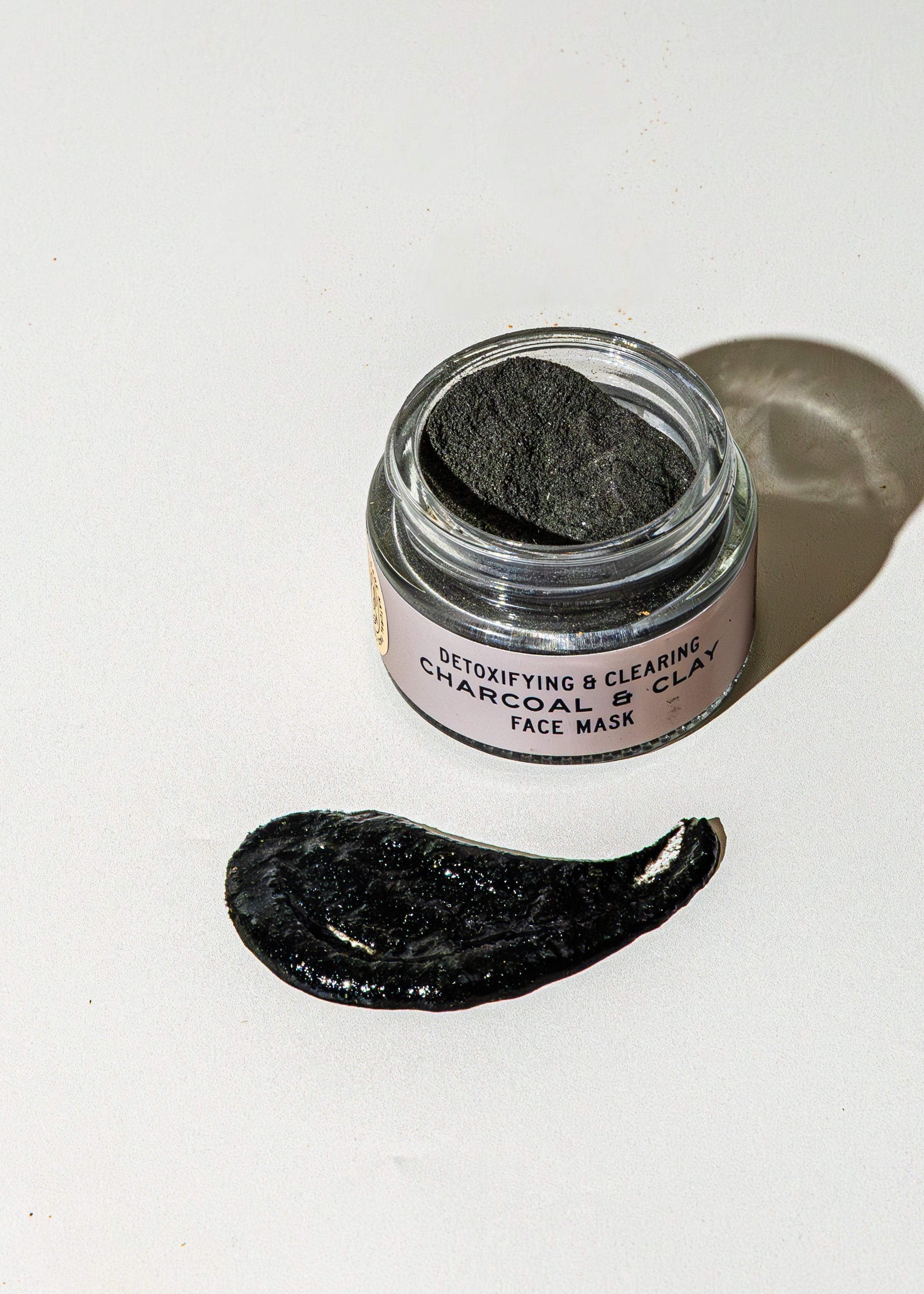 An open jar of Good Flower Farm’s TESTER — Charcoal & Clay Botanical Face Mask sits on a white surface, a dark smear in front. The label reads “Detoxifying & Clearing Charcoal Clay Face Mask.”.