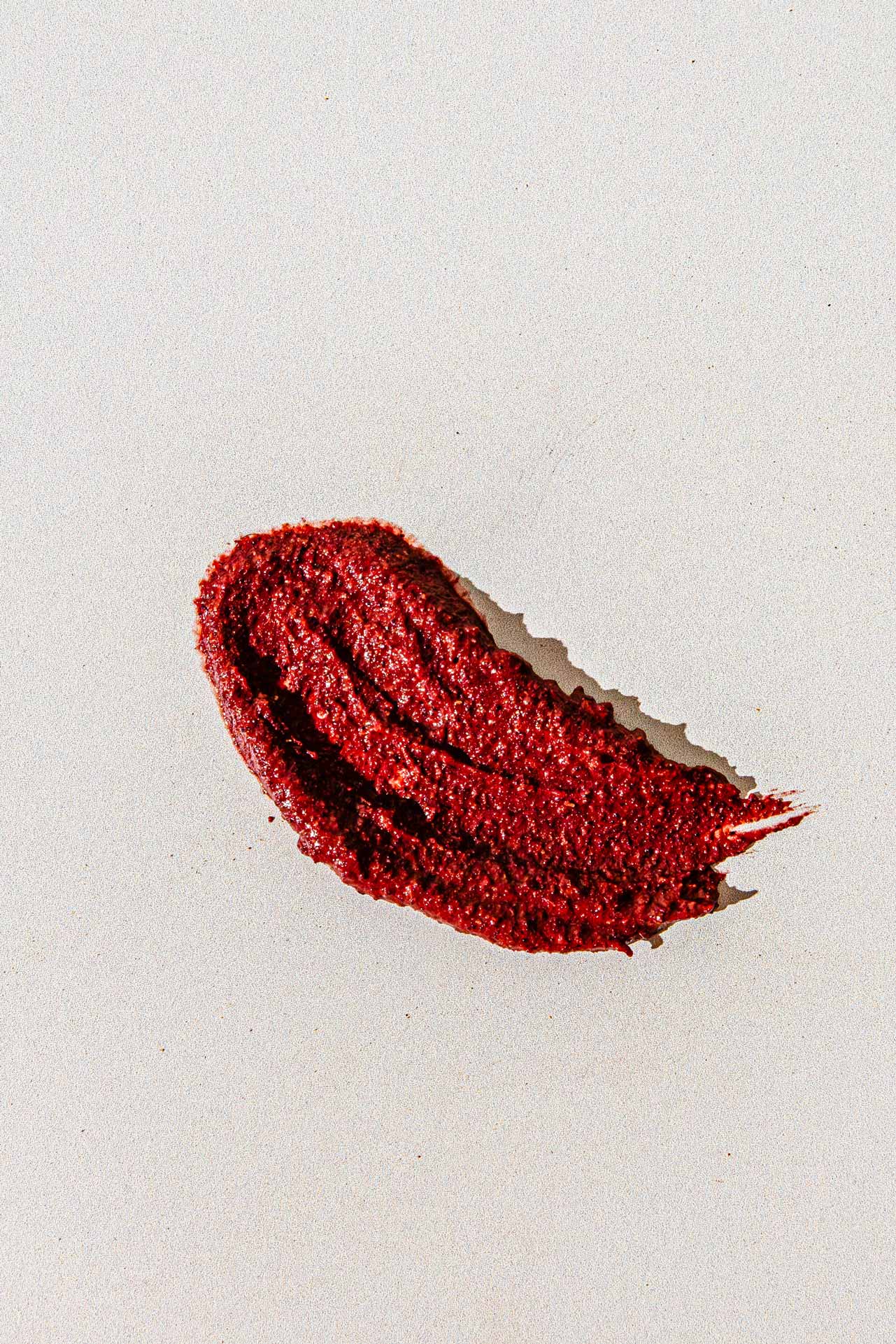 A smear of deep red Flowers & Rose Clay Botanical Face Mask by Good Flower Farm—reminiscent of rosehips—spreads across a pale, off-white background, showcasing textured brush strokes and a slightly uneven edge.