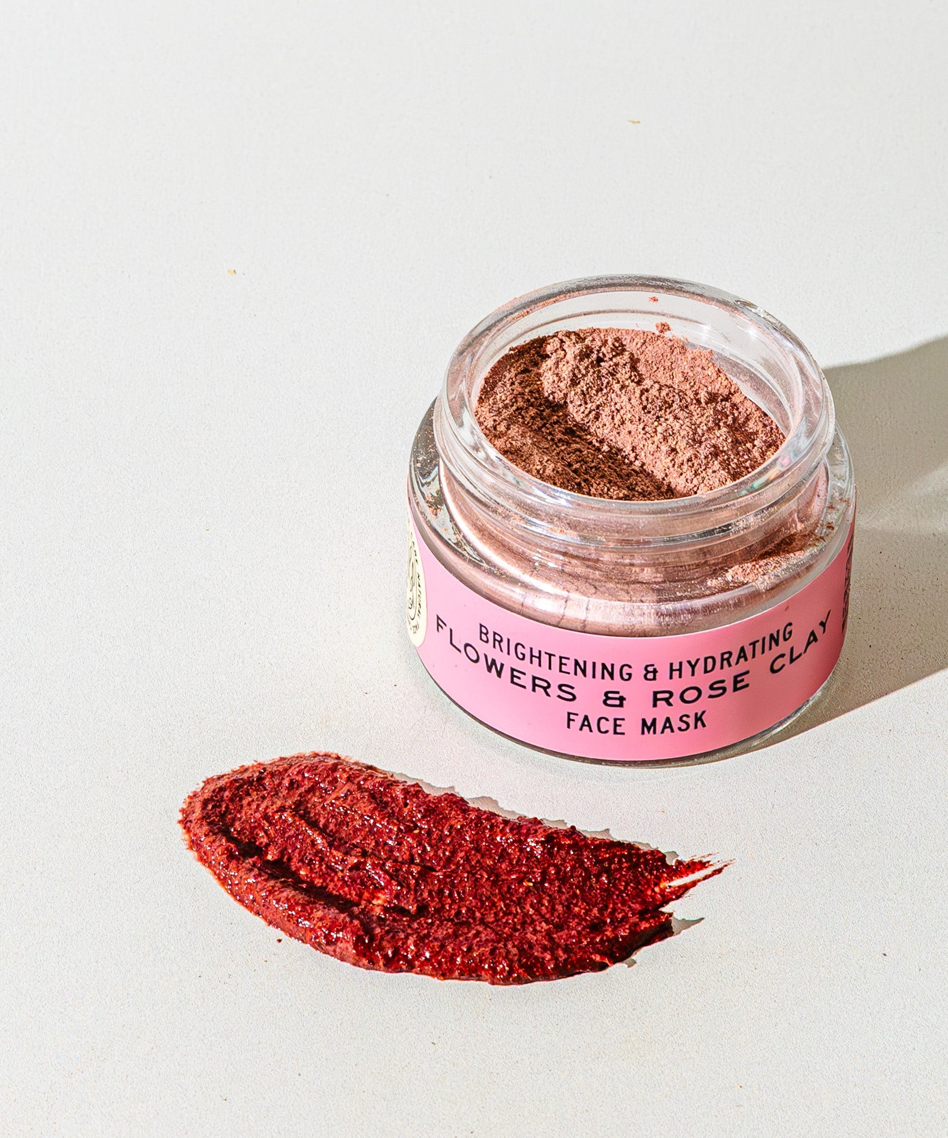 An open jar of TESTER — Flowers & Rose Clay Botanical Face Mask by Good Flower Farm rests on a light surface, showcasing its botanical formula with a swatch of the rosy clay mask displayed in front.