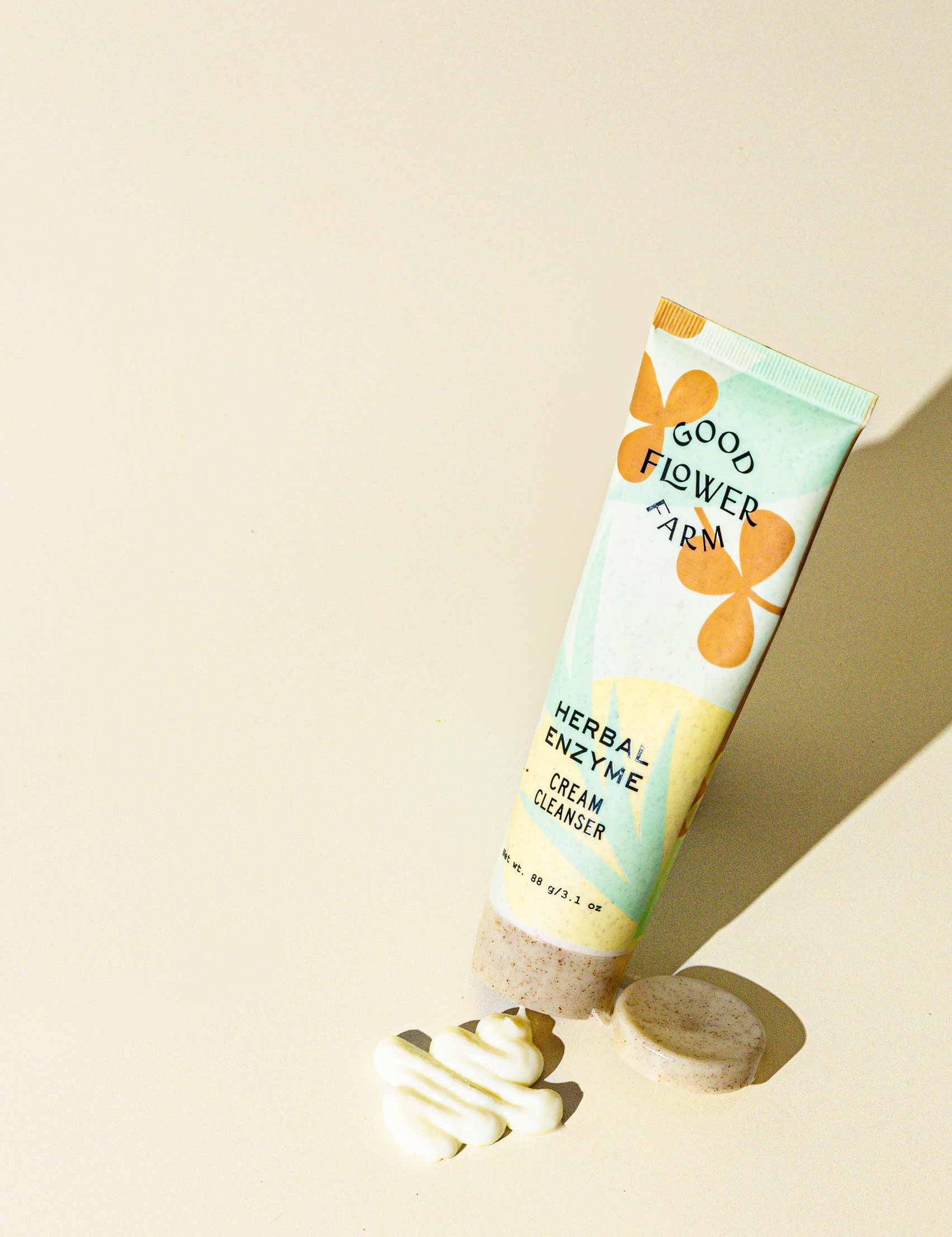 A tube of Good Flower Farm Herbal Enzyme Cream Cleanser & Makeup Remover stands upright on a light surface beside a round bar and a swirl of white cream, highlighted by bright natural lighting.