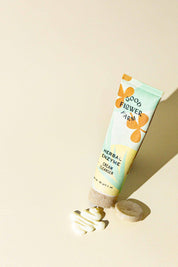 A tube of Good Flower Farm Herbal Enzyme Cream Cleanser & Makeup Remover stands upright on a light surface beside a round bar and a swirl of white cream, highlighted by bright natural lighting.