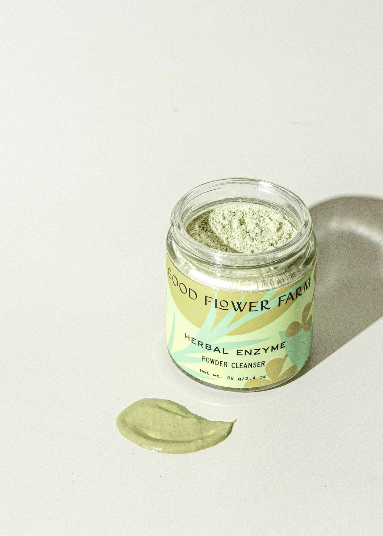 A jar of Good Flower Farm Herbal Enzyme Powder Facial Cleanser sits on a light surface, with some green powder—featuring papaya and pineapple enzymes—spread out in front of it.