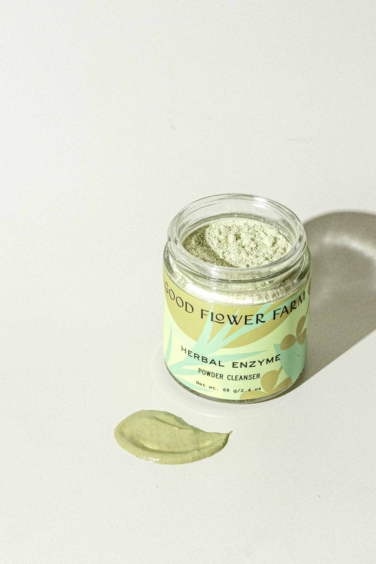 A jar of Good Flower Farm Herbal Enzyme Powder Facial Cleanser sits on a light surface, with some green powder—featuring papaya and pineapple enzymes—spread out in front of it.