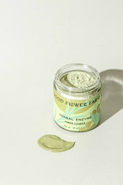 A jar of Good Flower Farm Herbal Enzyme Powder Facial Cleanser sits on a light surface, with some green powder—featuring papaya and pineapple enzymes—spread out in front of it.