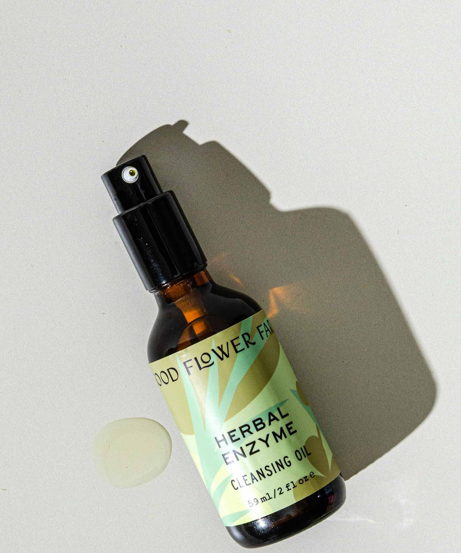 A brown glass spray bottle of Good Flower Farms Herbal Enzyme Cleansing Oil, featuring a green label with leaf graphics, rests on a light surface beside a small oil spill and casts a shadow, highlighting its purifying, papaya seed oil blend.