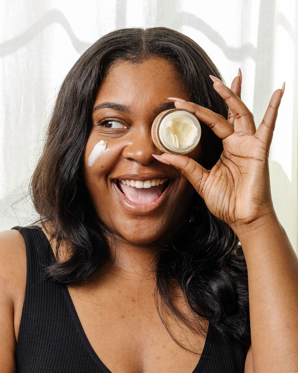 A smiling woman with long dark hair and a black top playfully holds a jar of Good Flower Farm Magic Mushroom Moisture Cream over one eye, with a dab on her cheek, posing against a light background.