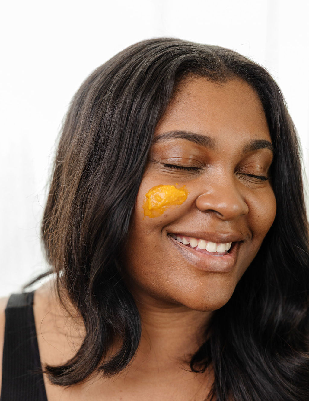 A smiling woman with closed eyes wears Good Flower Farm’s Golden Glow Beauty Balm on her cheek. With long dark hair and a black tank top, she radiates confidence against a light background.