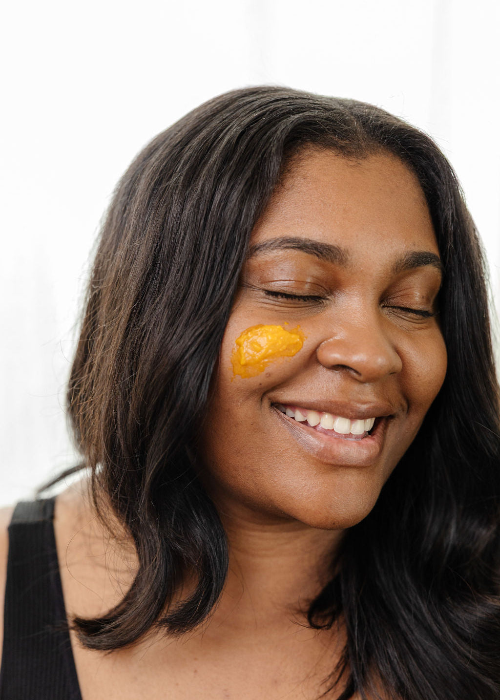 A smiling woman with closed eyes wears Good Flower Farm’s Golden Glow Beauty Balm on her cheek. With long dark hair and a black tank top, she radiates confidence against a light background.