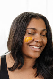 A smiling woman with closed eyes wears Good Flower Farm’s Golden Glow Beauty Balm on her cheek. With long dark hair and a black tank top, she radiates confidence against a light background.