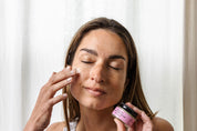 A woman with closed eyes applies Good Flower Farm Rose Geranium Face Cream to her cheek, holding a small jar with a pink label in front of a light curtain.