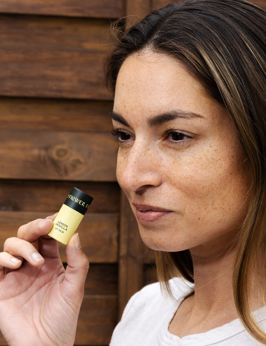 A woman with light brown hair and freckles holds a Good Flower Farm Lemon Vanilla Organic Lip Balm near her face, standing in front of a wooden background.