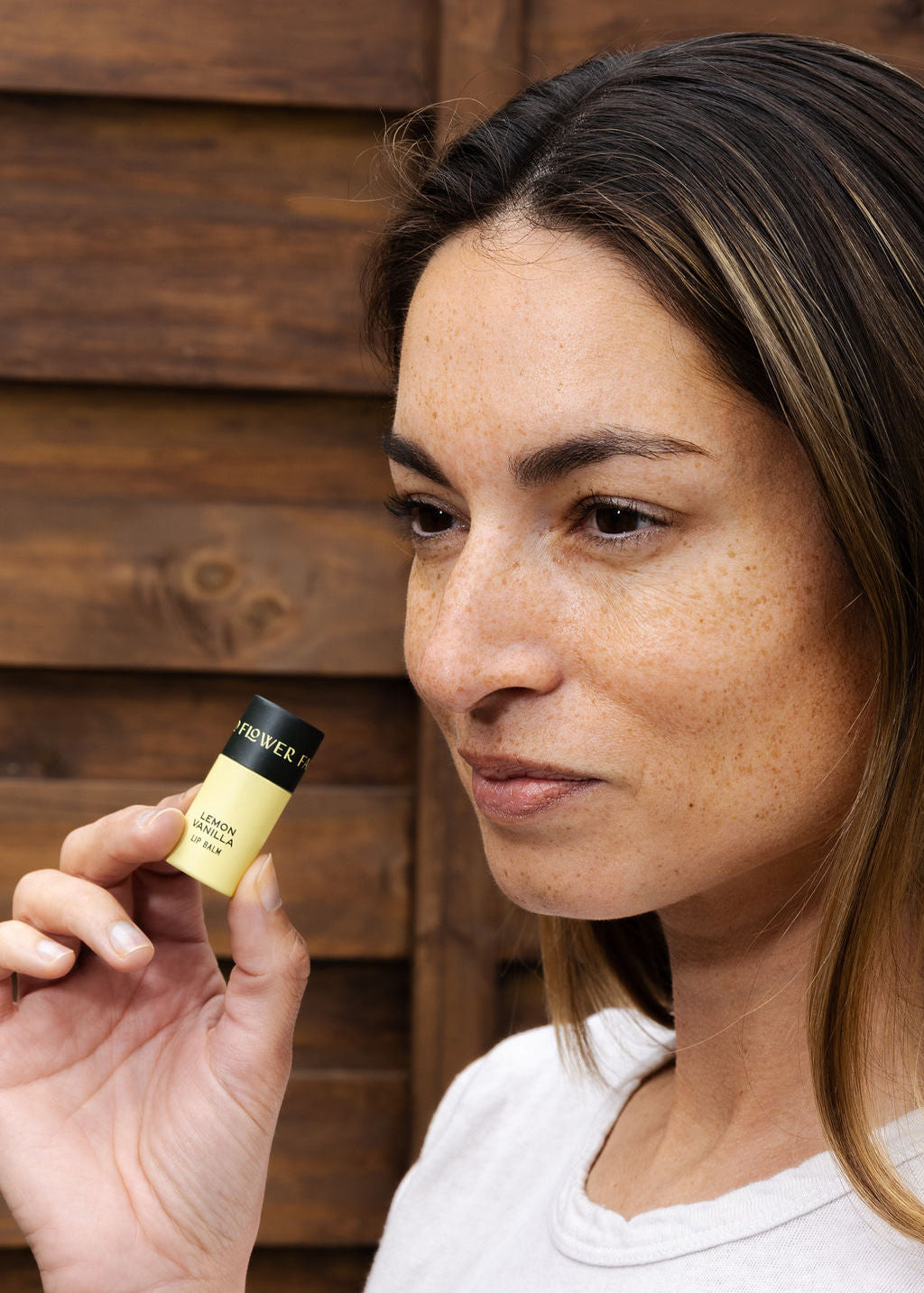 A woman with light brown hair and freckles holds a Good Flower Farm Lemon Vanilla Organic Lip Balm near her face, standing in front of a wooden background.