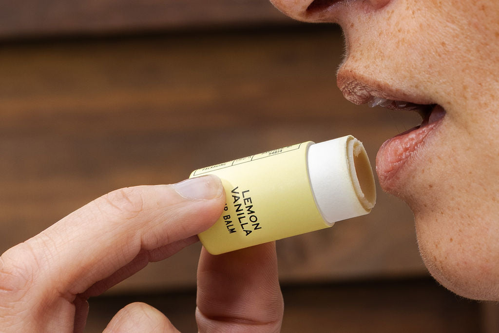 A person applies Good Flower Farm’s Lemon Vanilla Organic Lip Balm, holding the tube near their lips. The background features a soft, blurred wooden texture.