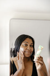 A woman smiles in front of a mirror as she applies Good Flower Farms Herbal Enzyme Cream Cleanser & Makeup Remover, infused with plant-based enzymes, to her face.