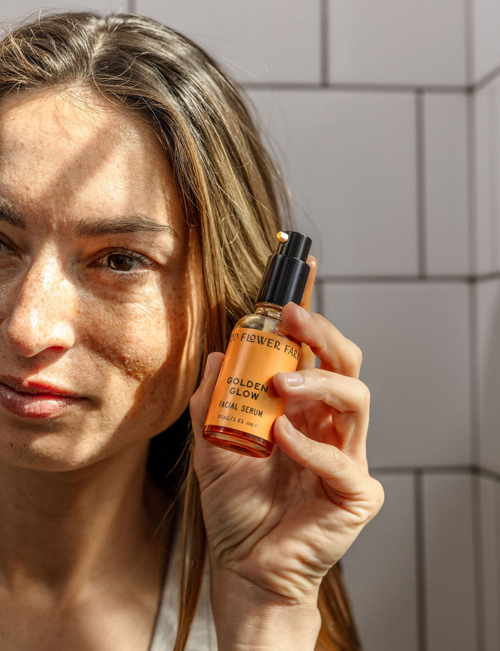 A woman with long brown hair holds up a bottle of Good Flower Farms Golden Glow Facial Serum in sunlight against a tiled wall, showcasing the serums promise of natural radiance and skin hydration.