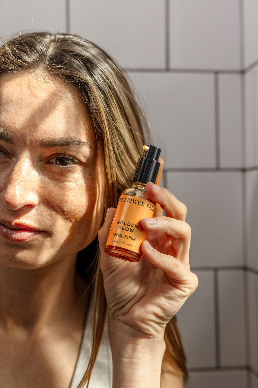 A woman with long brown hair holds up a bottle of Good Flower Farms Golden Glow Facial Serum in sunlight against a tiled wall, showcasing the serums promise of natural radiance and skin hydration.