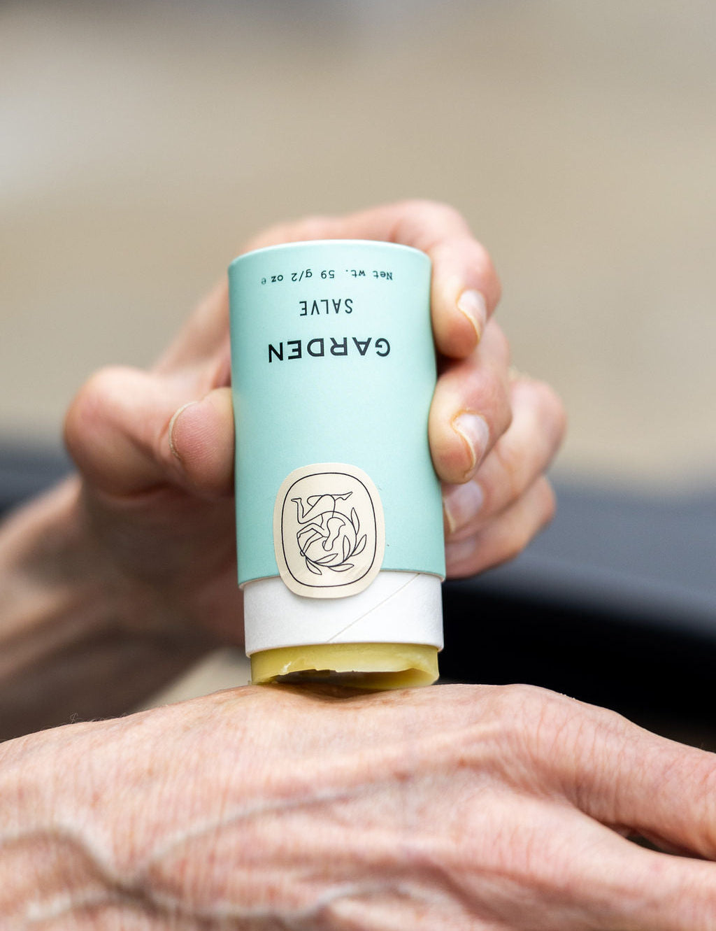 A person applies Good Flower Farms Garden Salve from a light blue tube with a beige cap and botanical logo to their hand; the salve is crafted with nourishing farm-grown herbs to soothe skin.