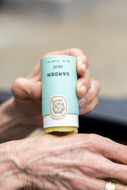A person applies Good Flower Farms Garden Salve from a light blue tube with a beige cap and botanical logo to their hand; the salve is crafted with nourishing farm-grown herbs to soothe skin.