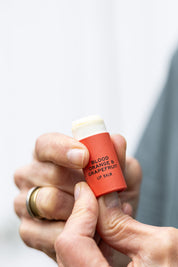 A person holds a Good Flower Farm Blood Orange & Grapefruit Organic Lip Balm with the cap off, showing the moisturizing balm. Against a light background, they wear a gold ring.