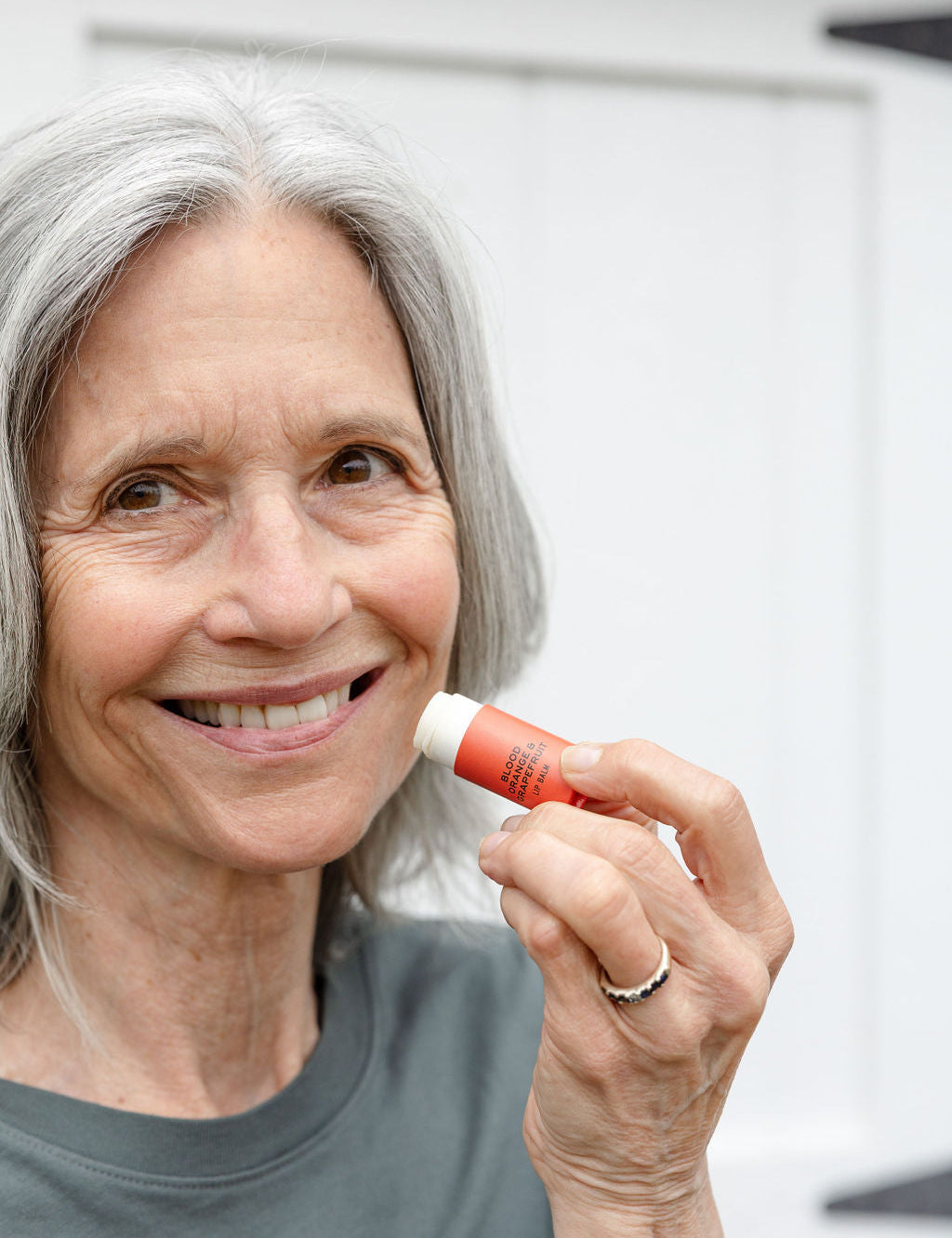 An older woman with gray hair smiles while holding Good Flower Farms Blood Orange & Grapefruit Organic Lip Balm near her lips, standing in front of a white background and wearing a gray shirt.