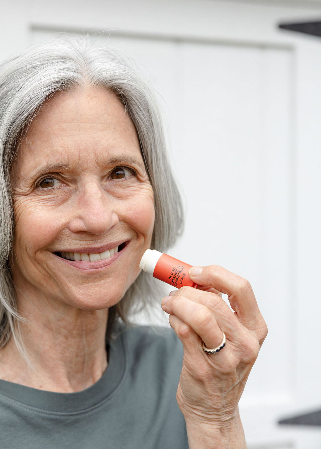 An older woman with gray hair smiles while holding Good Flower Farms Blood Orange & Grapefruit Organic Lip Balm near her lips, standing in front of a white background and wearing a gray shirt.