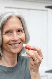 An older woman with gray hair smiles while holding Good Flower Farms Blood Orange & Grapefruit Organic Lip Balm near her lips, standing in front of a white background and wearing a gray shirt.