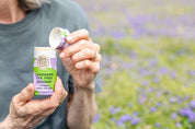 Someone in a gray shirt holds an open Good Flower Farm Lavender Tea Tree Deodorant Plastic-Free Tube, with a blurred field of purple flowers in the background.
