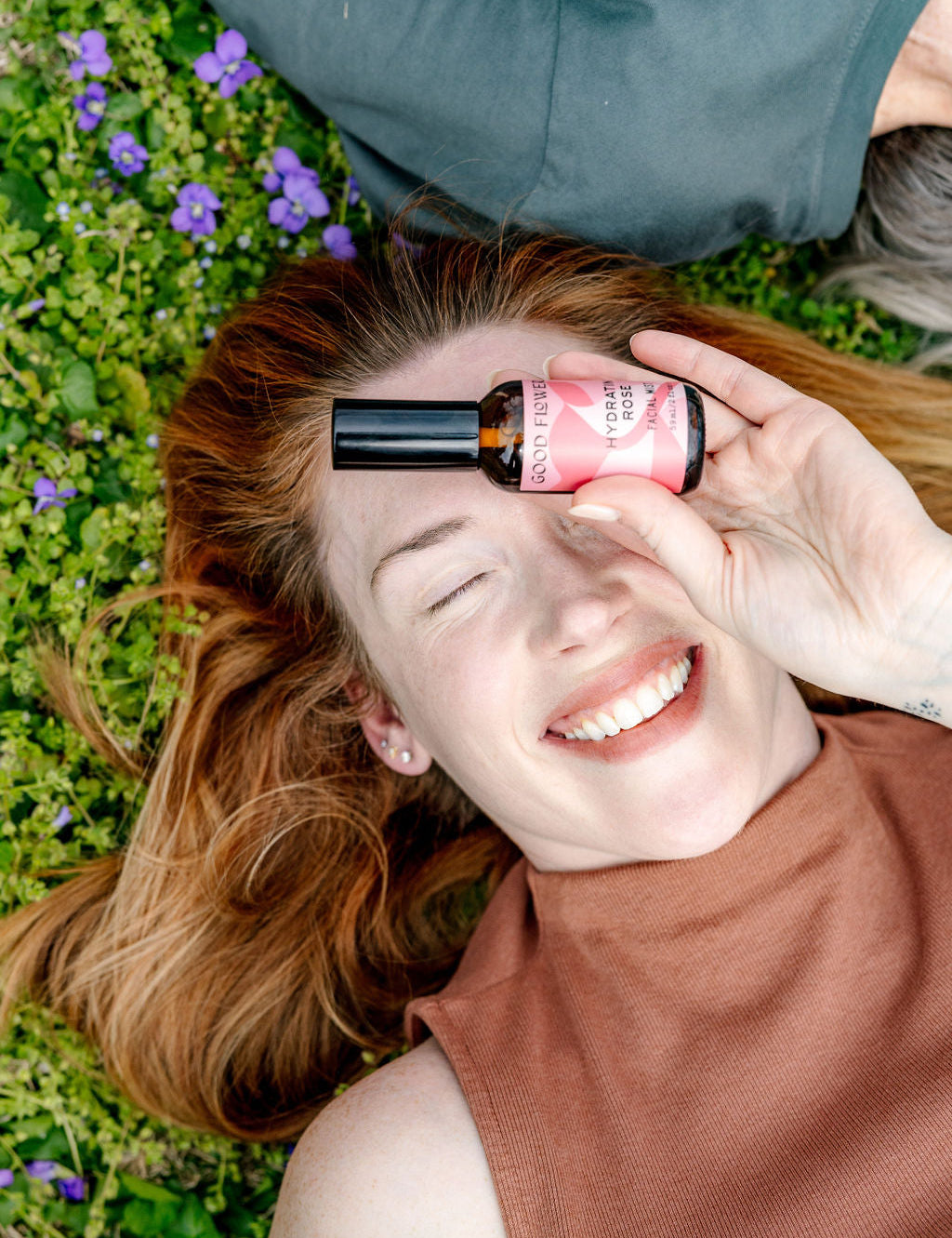 A joyful red-haired woman lies on grass with purple flowers, holding a pink bottle of Good Flower Farm Hydrating Rose Facial Mist before one eye. Wearing a sleeveless brown top, she enjoys refreshing hydration outdoors.