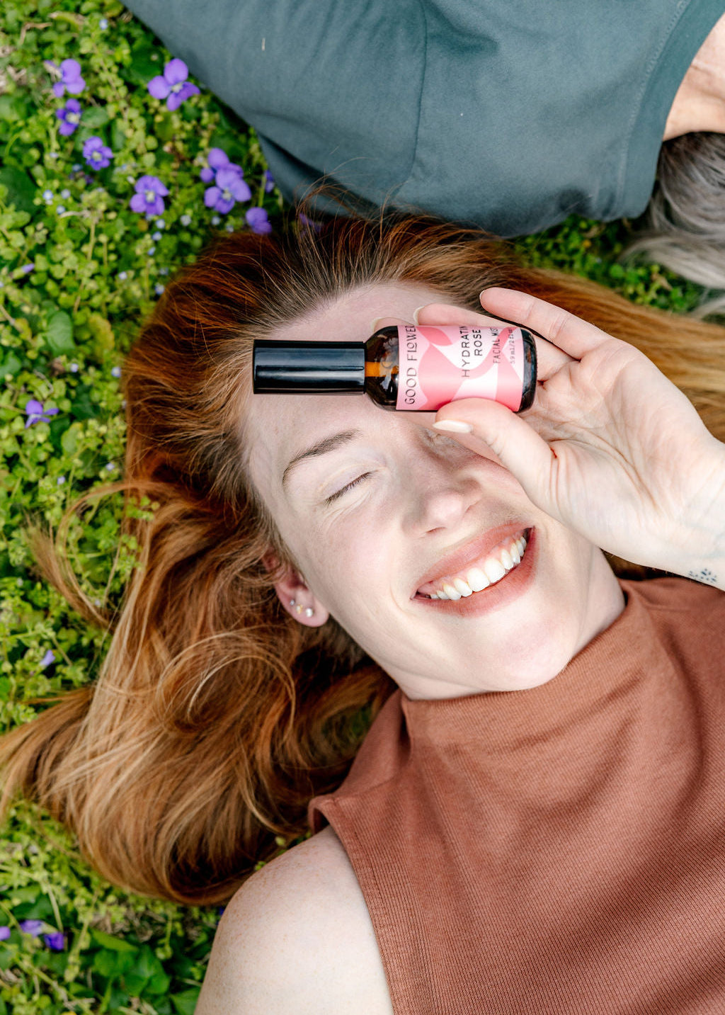 A joyful red-haired woman lies on grass with purple flowers, holding a pink bottle of Good Flower Farm Hydrating Rose Facial Mist before one eye. Wearing a sleeveless brown top, she enjoys refreshing hydration outdoors.