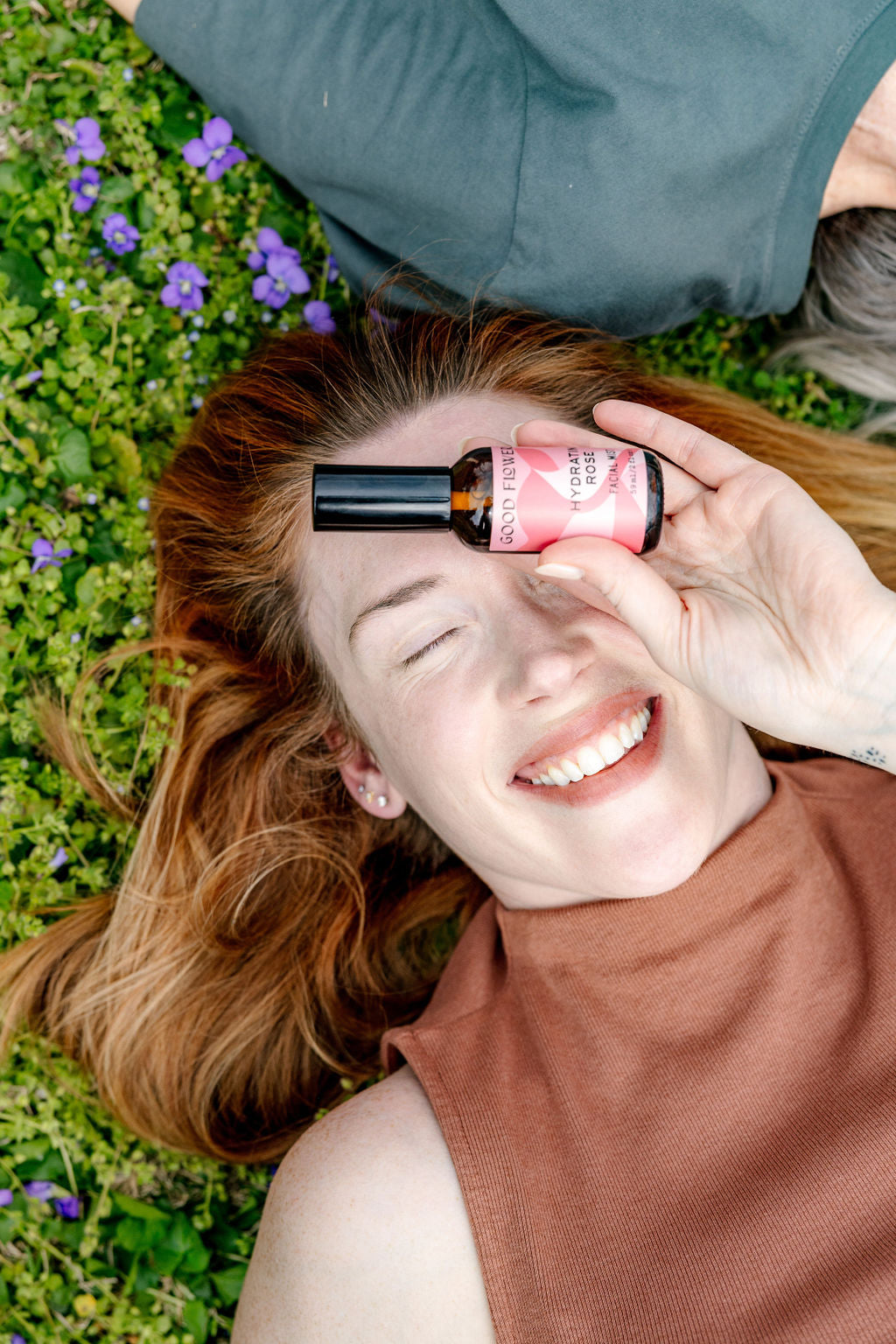 A joyful red-haired woman lies on grass with purple flowers, holding a pink bottle of Good Flower Farm Hydrating Rose Facial Mist before one eye. Wearing a sleeveless brown top, she enjoys refreshing hydration outdoors.