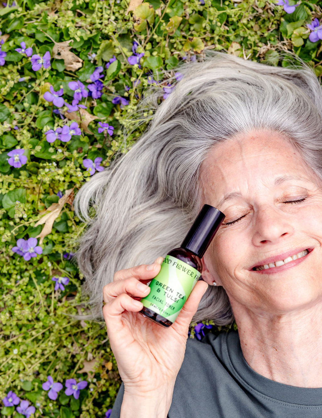 An older woman with gray hair lies on grass with purple flowers, smiling with eyes closed, holding a green bottle labeled Green Tea & Tulsi Facial Toner by Good Flower Farm—a soothing blend for your skin.