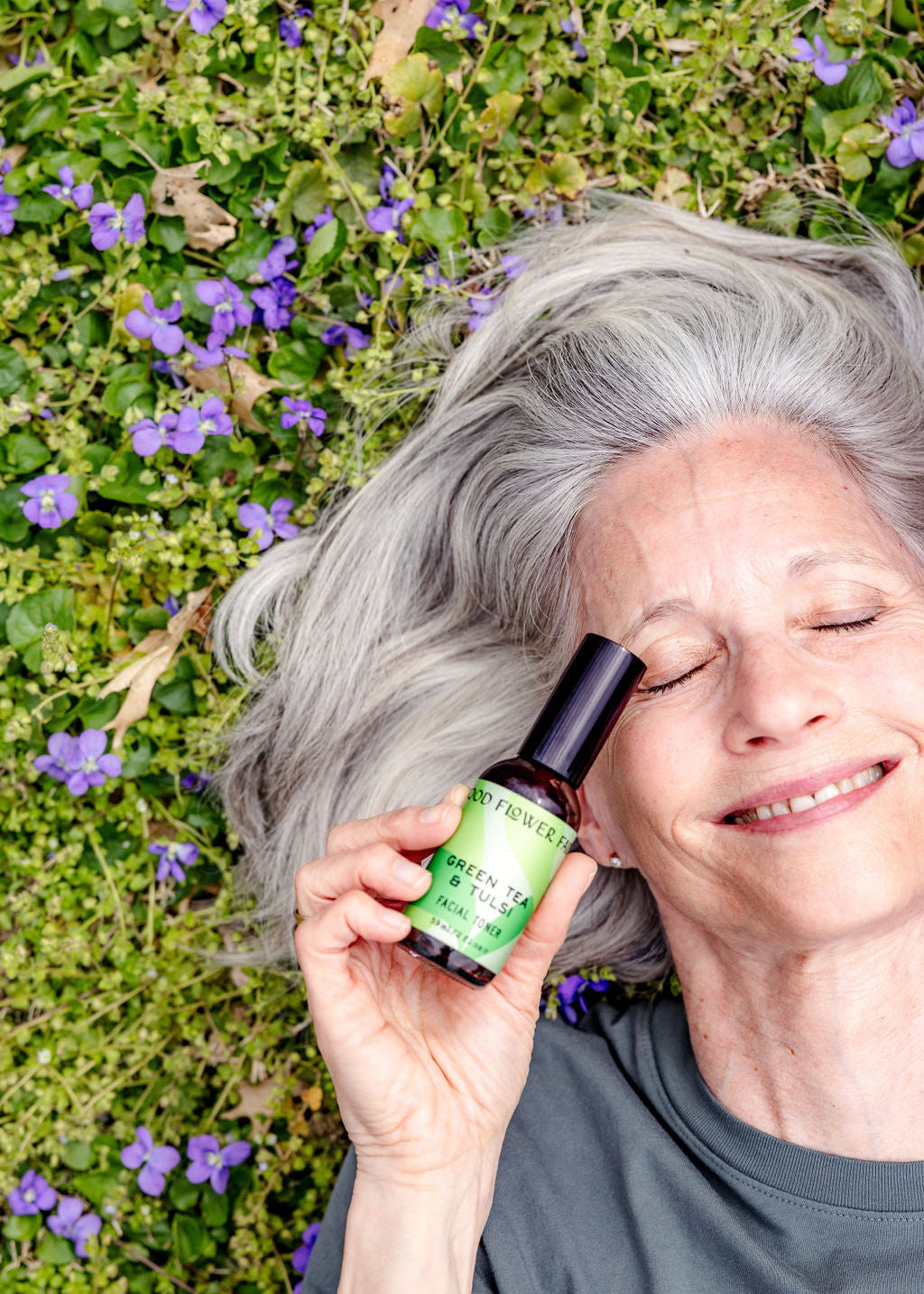 An older woman with gray hair lies on grass with purple flowers, smiling with eyes closed, holding a green bottle labeled Green Tea & Tulsi Facial Toner by Good Flower Farm—a soothing blend for your skin.