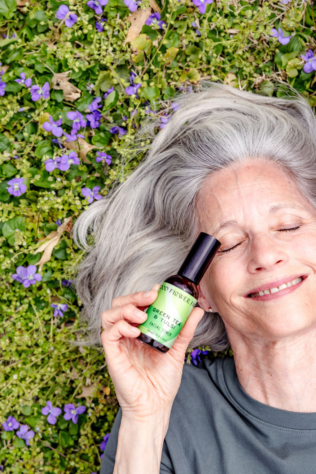 An older woman with gray hair lies on grass with purple flowers, smiling with eyes closed, holding a green bottle labeled Green Tea & Tulsi Facial Toner by Good Flower Farm—a soothing blend for your skin.