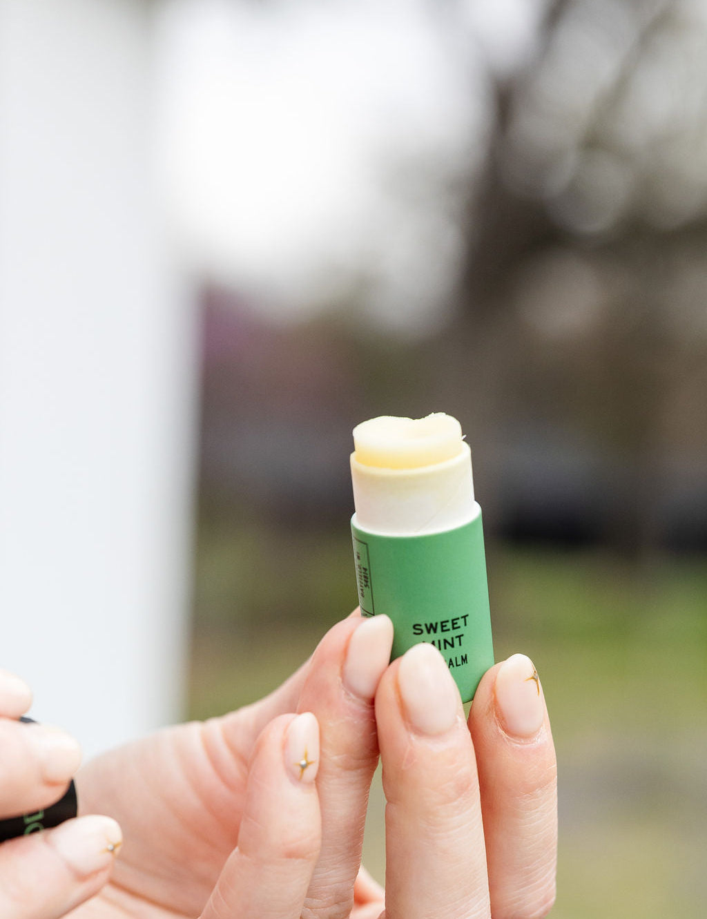 With manicured nails, someone holds an open tube of Good Flower Farm Sweet Mint Organic Lip Balm outdoors, with trees and grass softly blurred in the background.