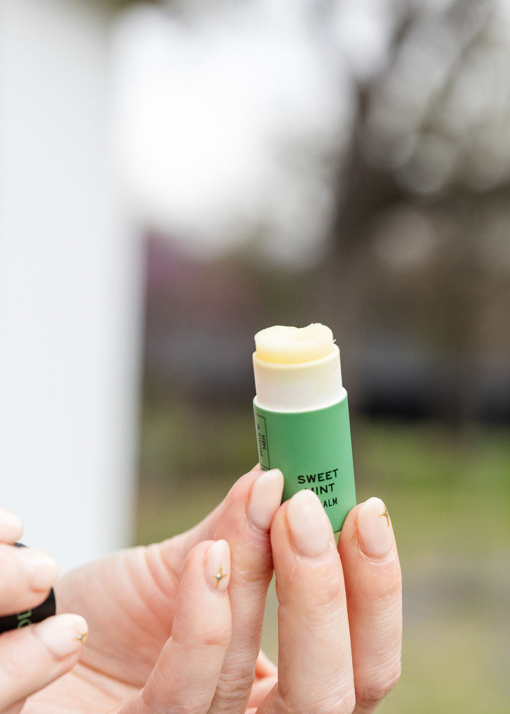 With manicured nails, someone holds an open tube of Good Flower Farm Sweet Mint Organic Lip Balm outdoors, with trees and grass softly blurred in the background.