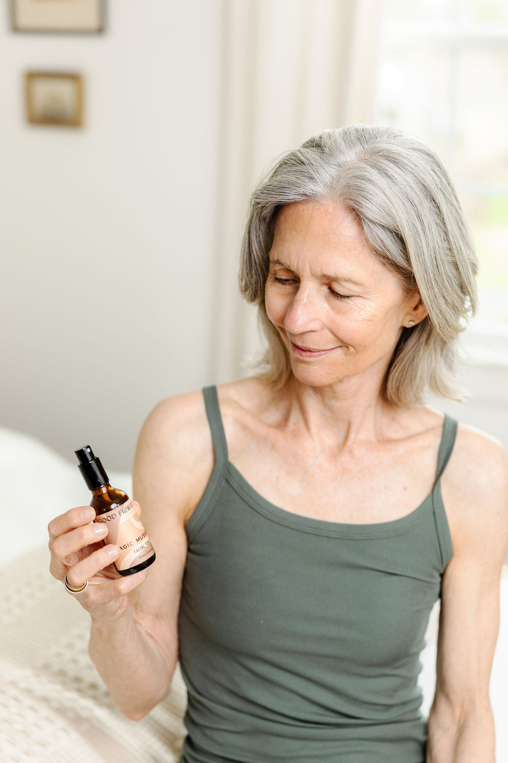 A smiling older woman with gray hair in a green tank top sits indoors, admiring a Good Flower Farm Magic Mushroom Facial Toner bottle—enjoying the ultimate skin hydration it provides.