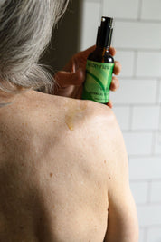 A person with gray hair applies Good Flower Farms Tulsi Botanical Body Oil from a green-labeled bottle onto their bare shoulder, a drop glistening on their skin against a white tiled wall backdrop.