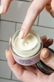 A person dips a finger into an open jar of Good Flower Farms Lavender Face Cream, revealing its calendula-infused, nourishing formula over a tiled surface.