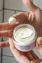 A hand holds an open jar of Good Flower Farm Lavender Face Cream, while the other hand scoops out a bit of the moisturizer. The product label is clearly visible.