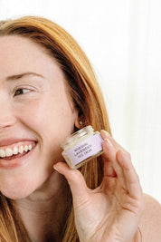 A smiling woman with long red hair holds a jar of Good Flower Farm’s Lavender Face Cream near her face, showing off its soothing and hydrating lavender formula.