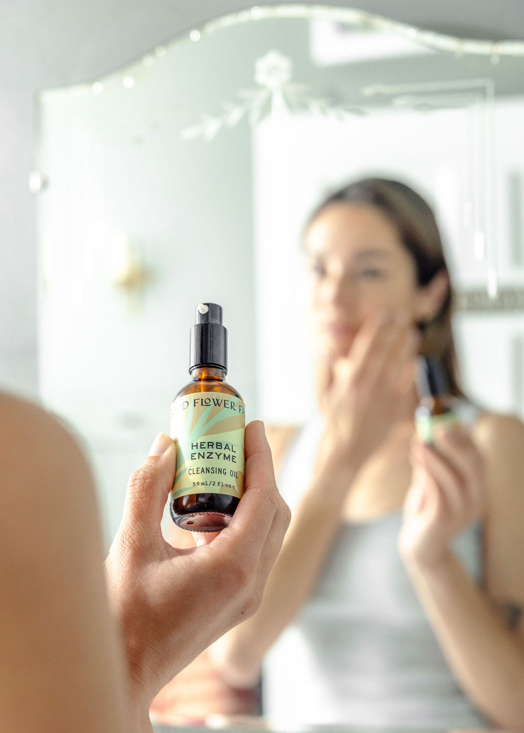 A person holds a bottle of Good Flower Farm Herbal Enzyme Cleansing Oil in focus, while their blurred reflection applies the product to their face in front of a mirror.