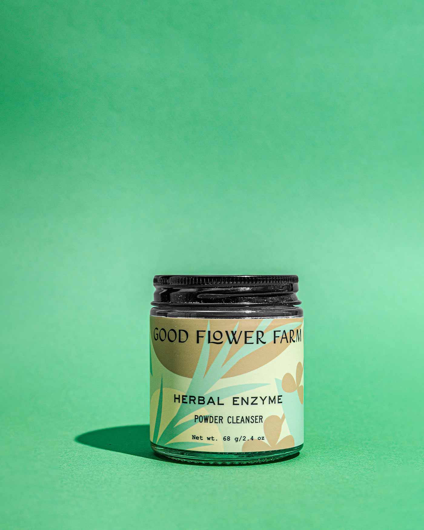 A jar of Good Flower Farm Herbal Enzyme Powder Facial Cleanser, featuring botanical illustrations and earthy colors on the label, is displayed on a green surface with a green gradient background.