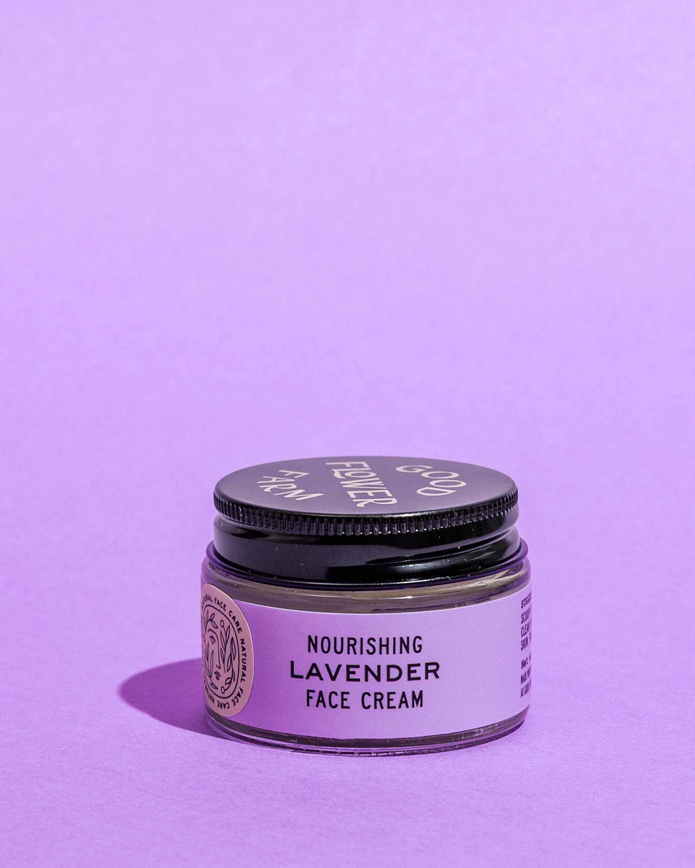 A small glass jar of Good Flower Farm Lavender Face Cream, made with lavender hydrosol, sits on a light purple surface against a matching background. The jar has a black lid and a lavender label.