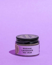 A small glass jar of Good Flower Farm Lavender Face Cream, made with lavender hydrosol, sits on a light purple surface against a matching background. The jar has a black lid and a lavender label.