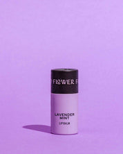 A lavender-colored biodegradable tube of Good Flower Farm’s Lavender Mint Organic Lip Balm stands upright on a matching lavender background, casting a left shadow. This soothing balm is enriched with nourishing coconut oil.