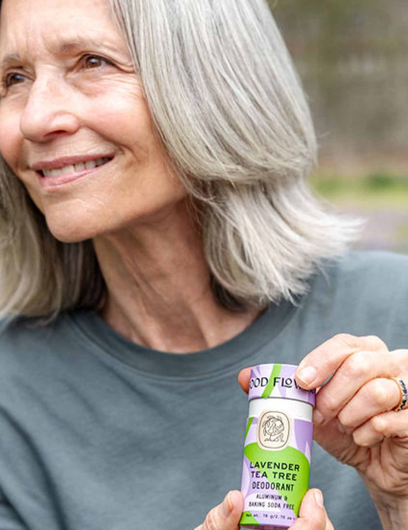 An older woman with gray hair smiles outdoors while holding a purple and green tube of Good Flower Farm Lavender Tea Tree Deodorant in a plastic-free tube.